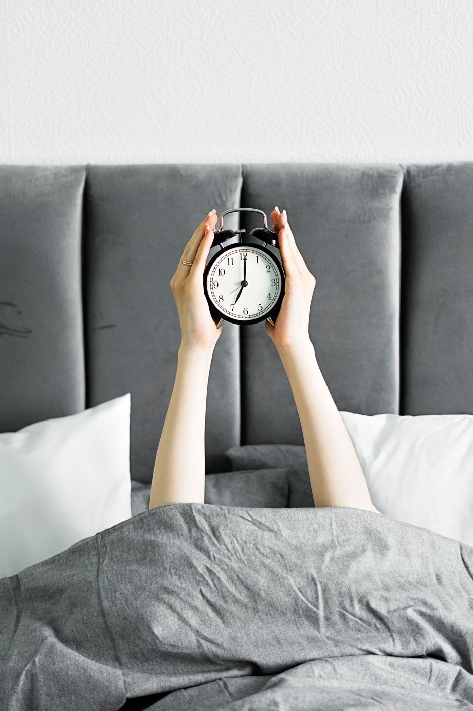 A hand holding a small analog alarm clock beside a bed